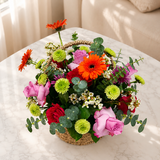 A vibrant flower arrangement in a woven basket, featuring orange gerbera daisies, pink roses, red carnations, lime green chrysanthemums, and eucalyptus leaves. fresh flowers delivery Dubai