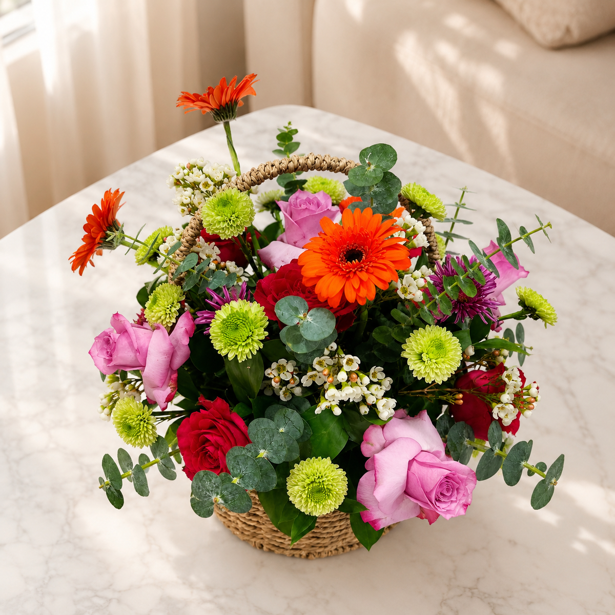 A vibrant flower arrangement in a woven basket, featuring orange gerbera daisies, pink roses, red carnations, lime green chrysanthemums, and eucalyptus leaves. fresh flowers delivery Dubai