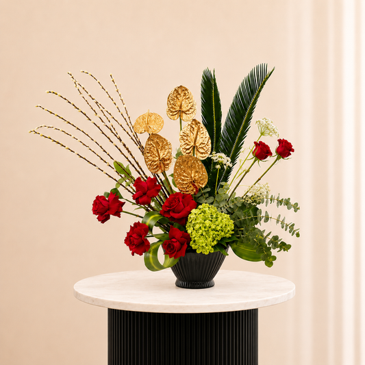 A floral arrangement featuring red roses, green hydrangea, red palm leaves, and gold anthuriums in a black ribbed vase, fresh flowers delivery Dubai.